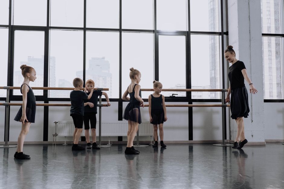 Young children learning ballet in a dance studio with floor-to-ceiling windows and a ballet instructor.